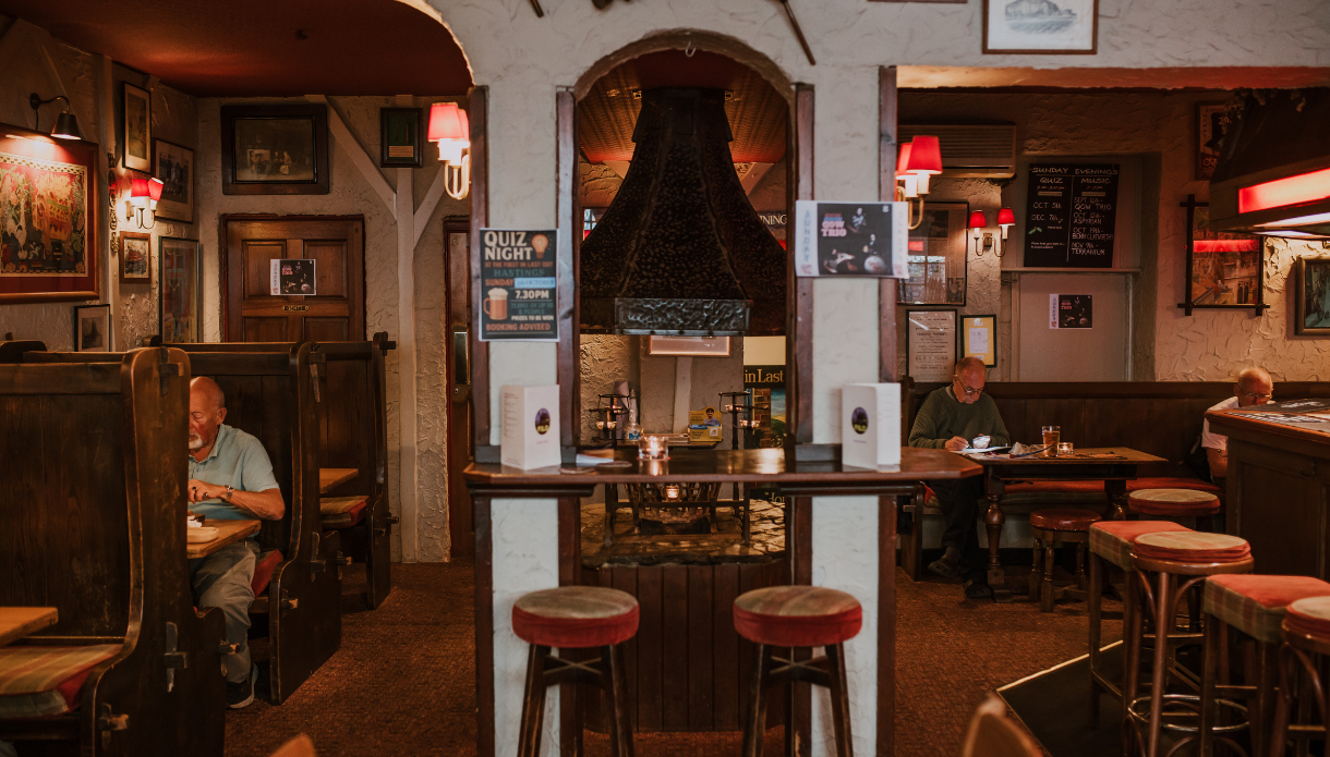 Cosy traditional pub with brown wood seating and a carpeted floor. In the middle, a black metal fireplace hood anchors the room, with two tall stools at a small counter. Soft red lights cast a warm glow on picture-filled walls and handwritten specials boards. Several guests dine quietly in booths and at small round tables. Overall mood is calm and intimate.